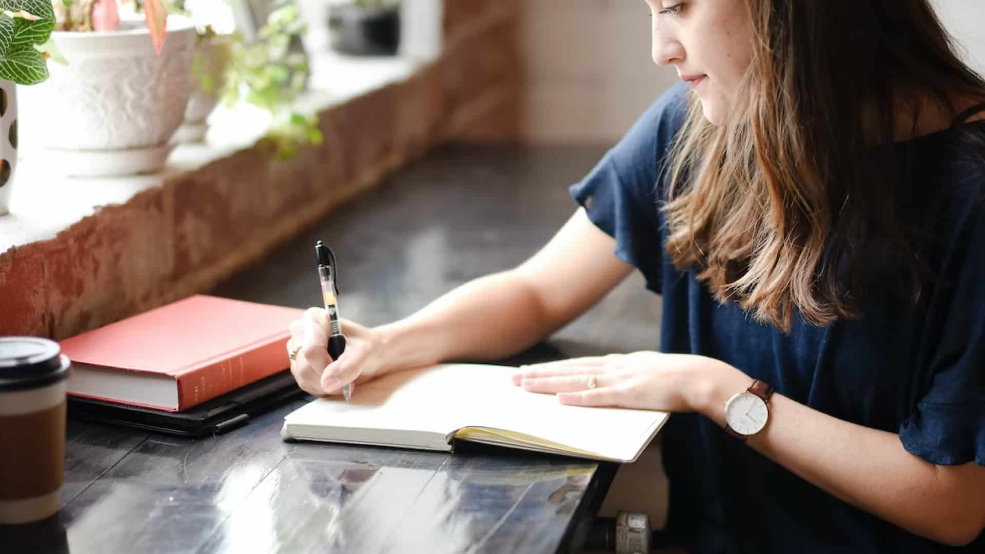Woman writing notes in notebook sitting a desk with coffee and other books
